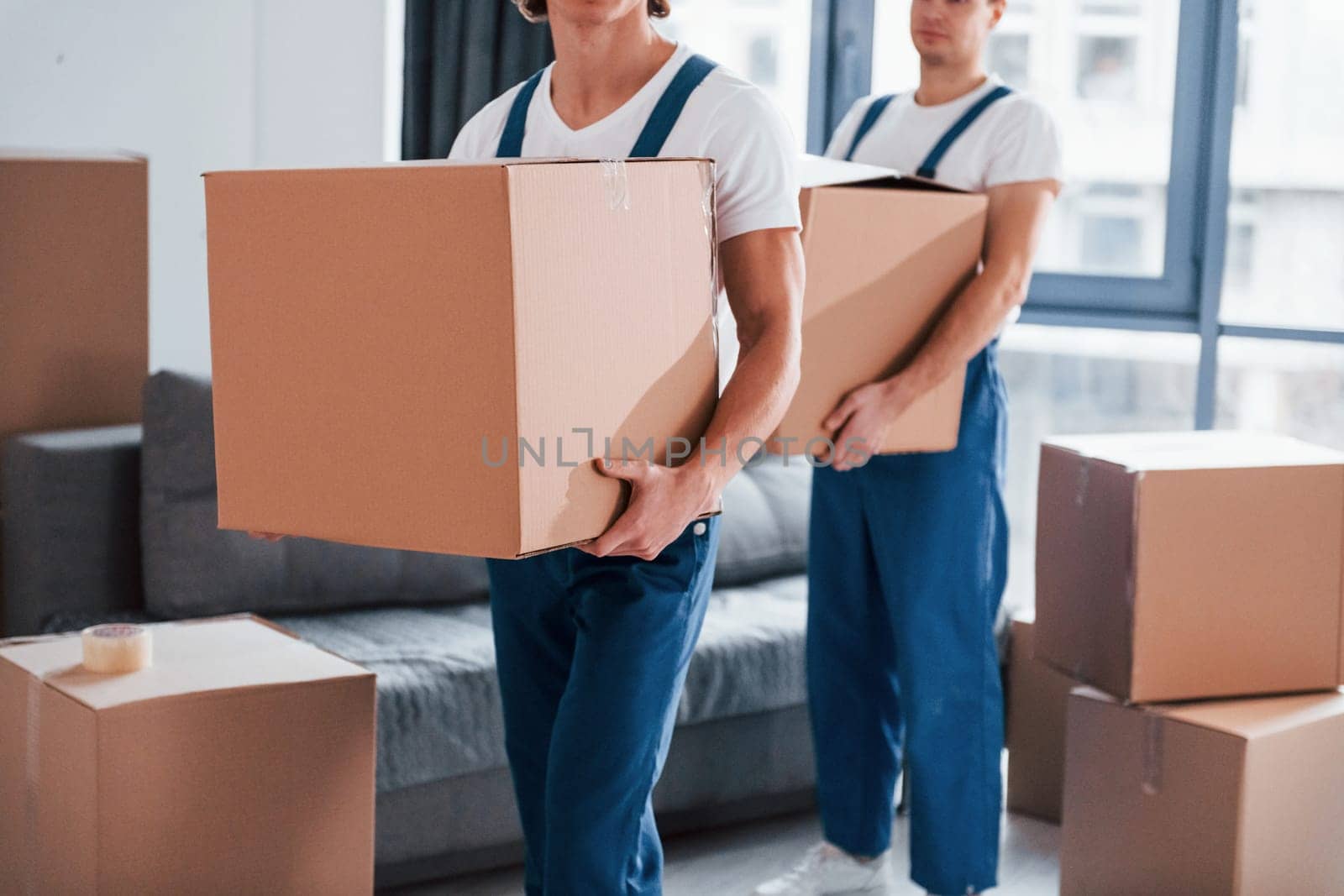 With boxes in hands. Two young movers in blue uniform working indoors in the room.
