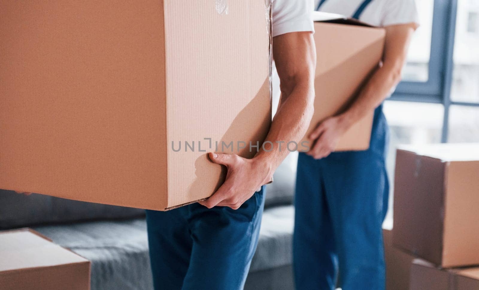 With boxes in hands. Two young movers in blue uniform working indoors in the room.