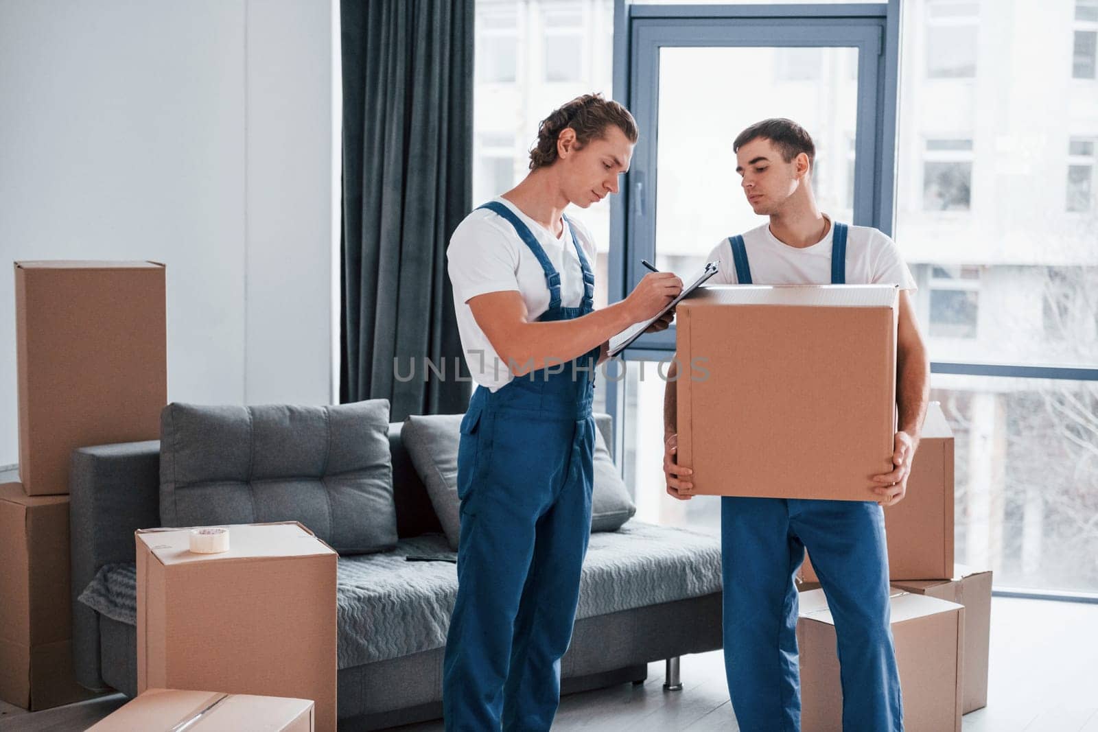 Holds notepad with document. Two young movers in blue uniform working indoors in the room.