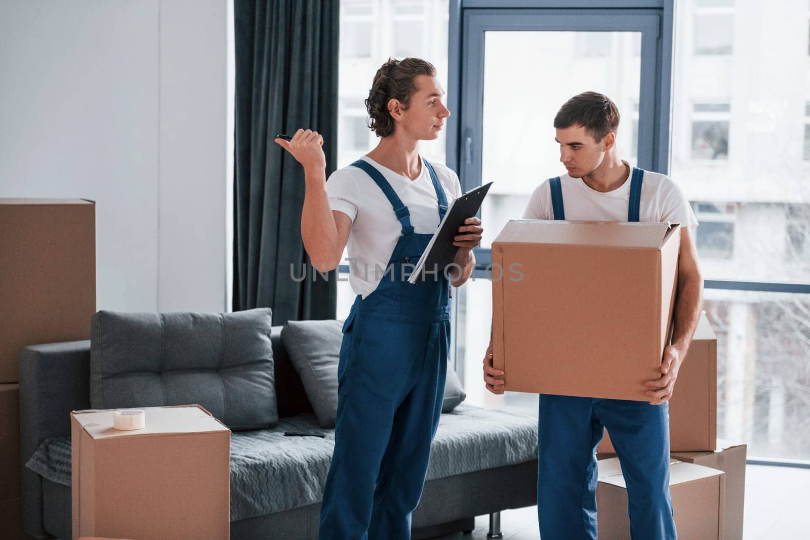 Holds notepad with document. Two young movers in blue uniform working indoors in the room.