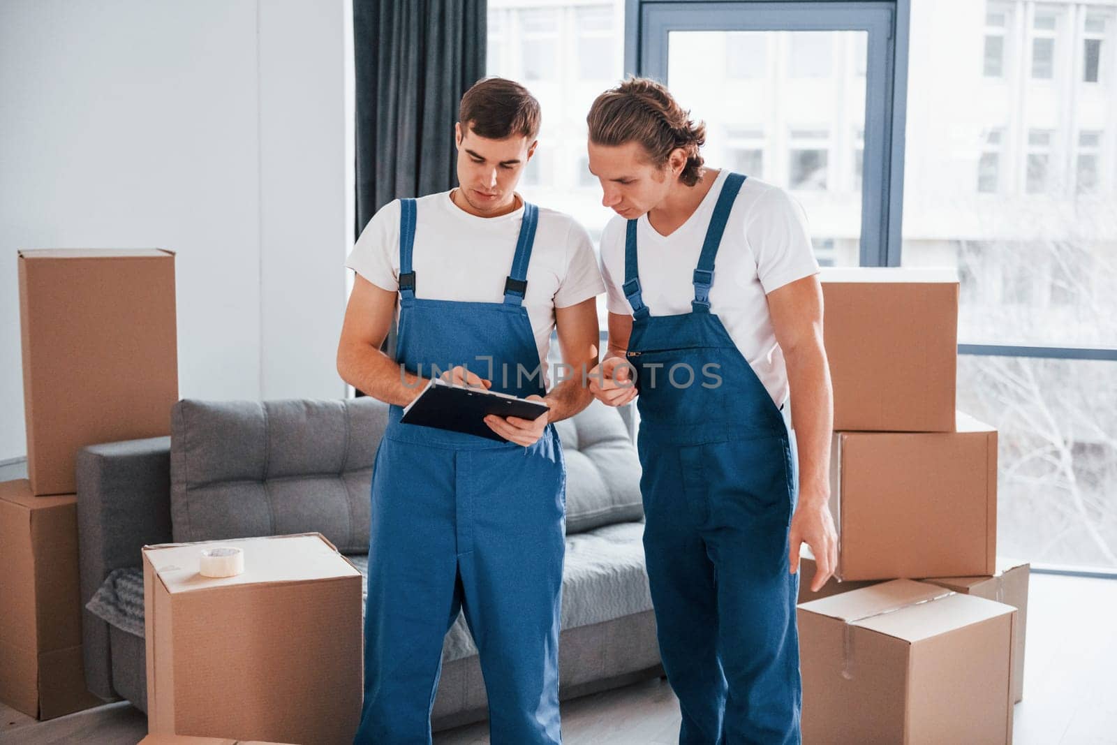 Holds notepad with document. Two young movers in blue uniform working indoors in the room.
