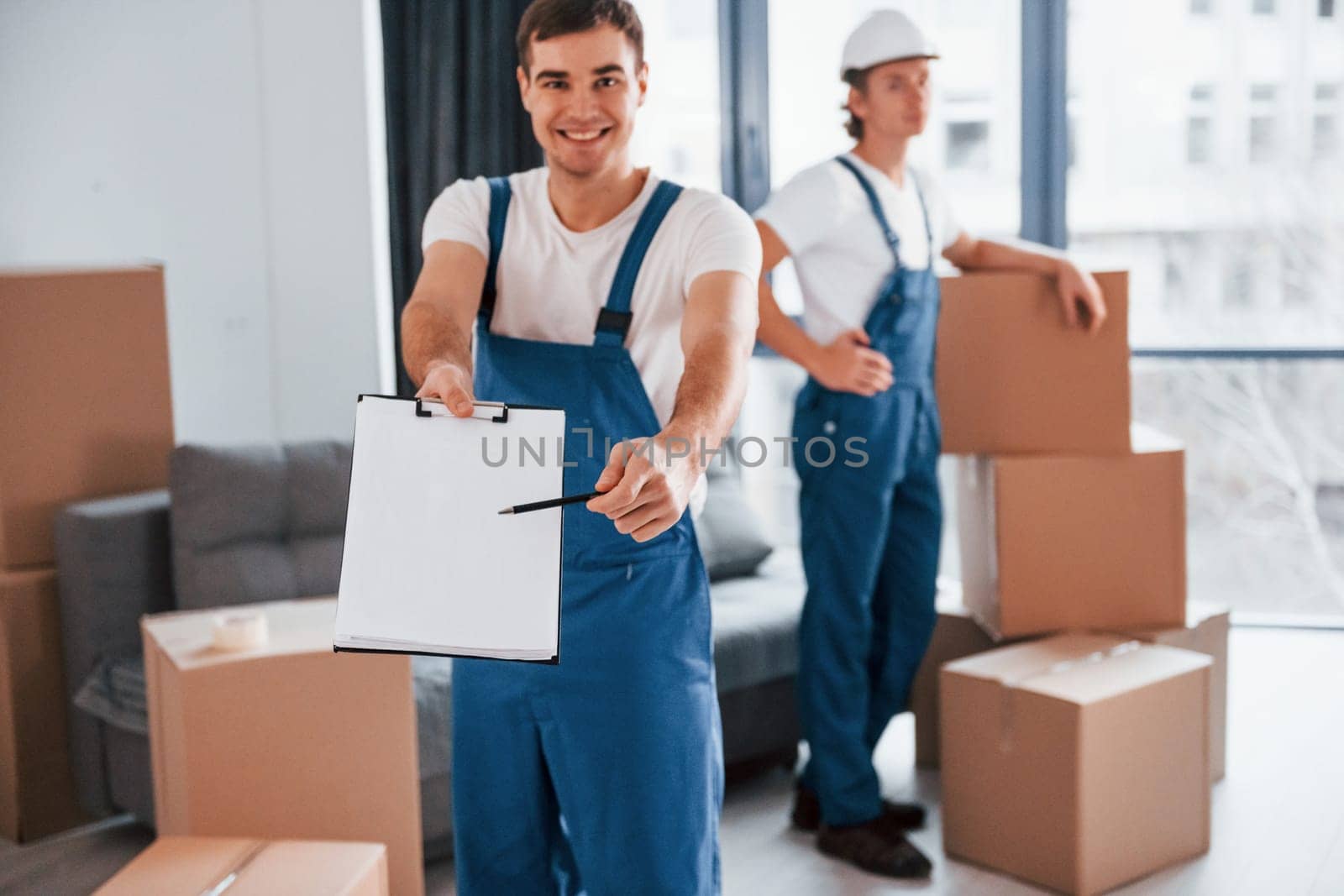 Document to sign. Two young movers in blue uniform working indoors in the room.