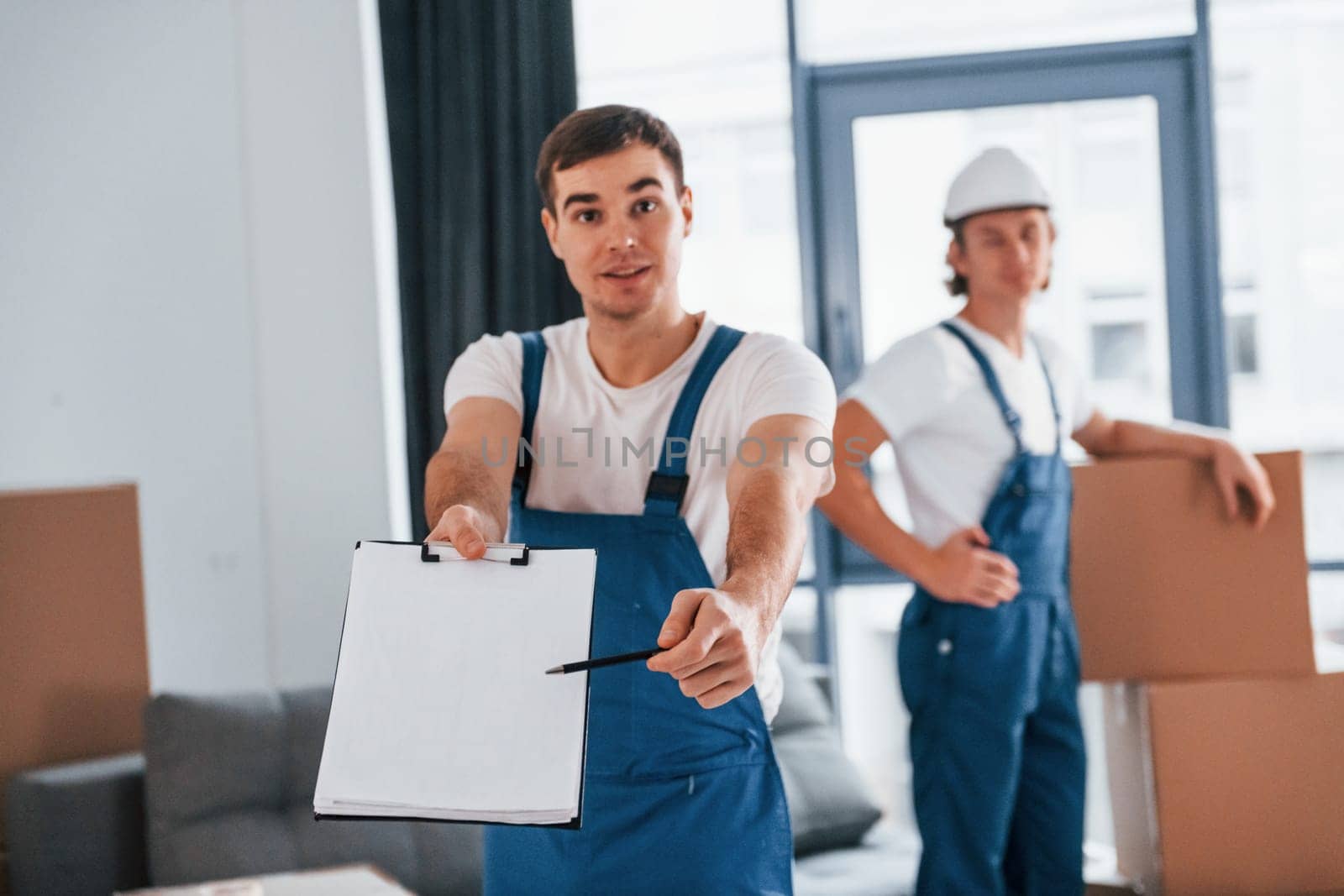Document to sign. Two young movers in blue uniform working indoors in the room.