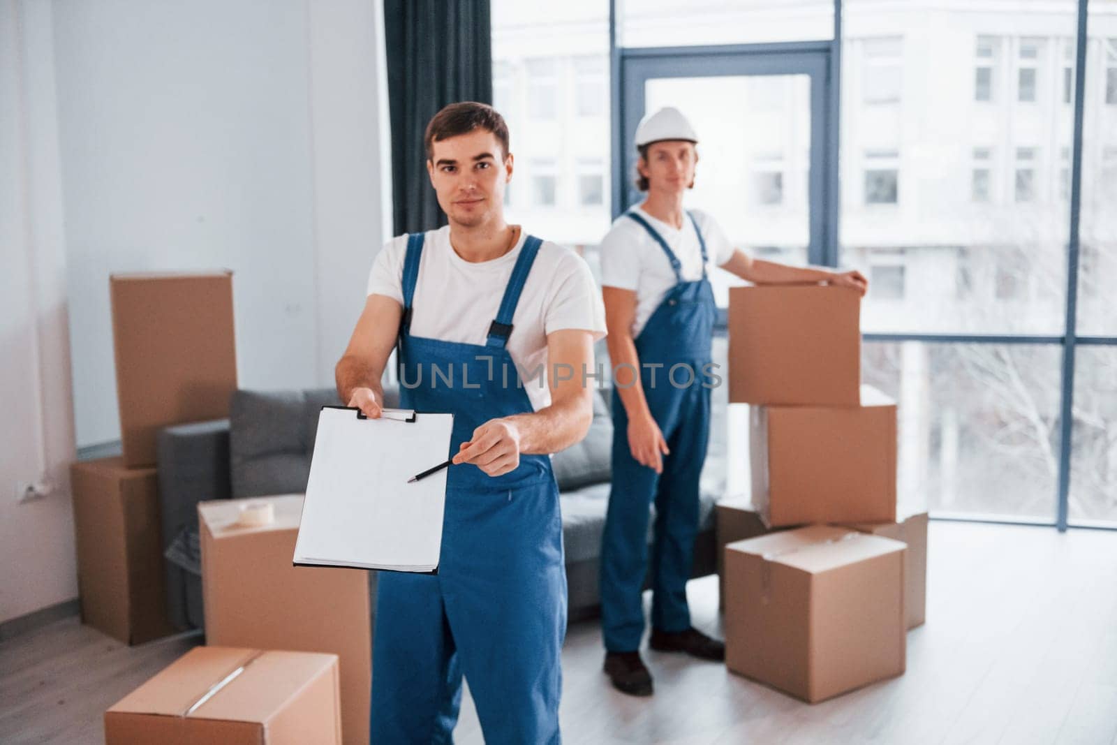 Document to sign. Two young movers in blue uniform working indoors in the room.