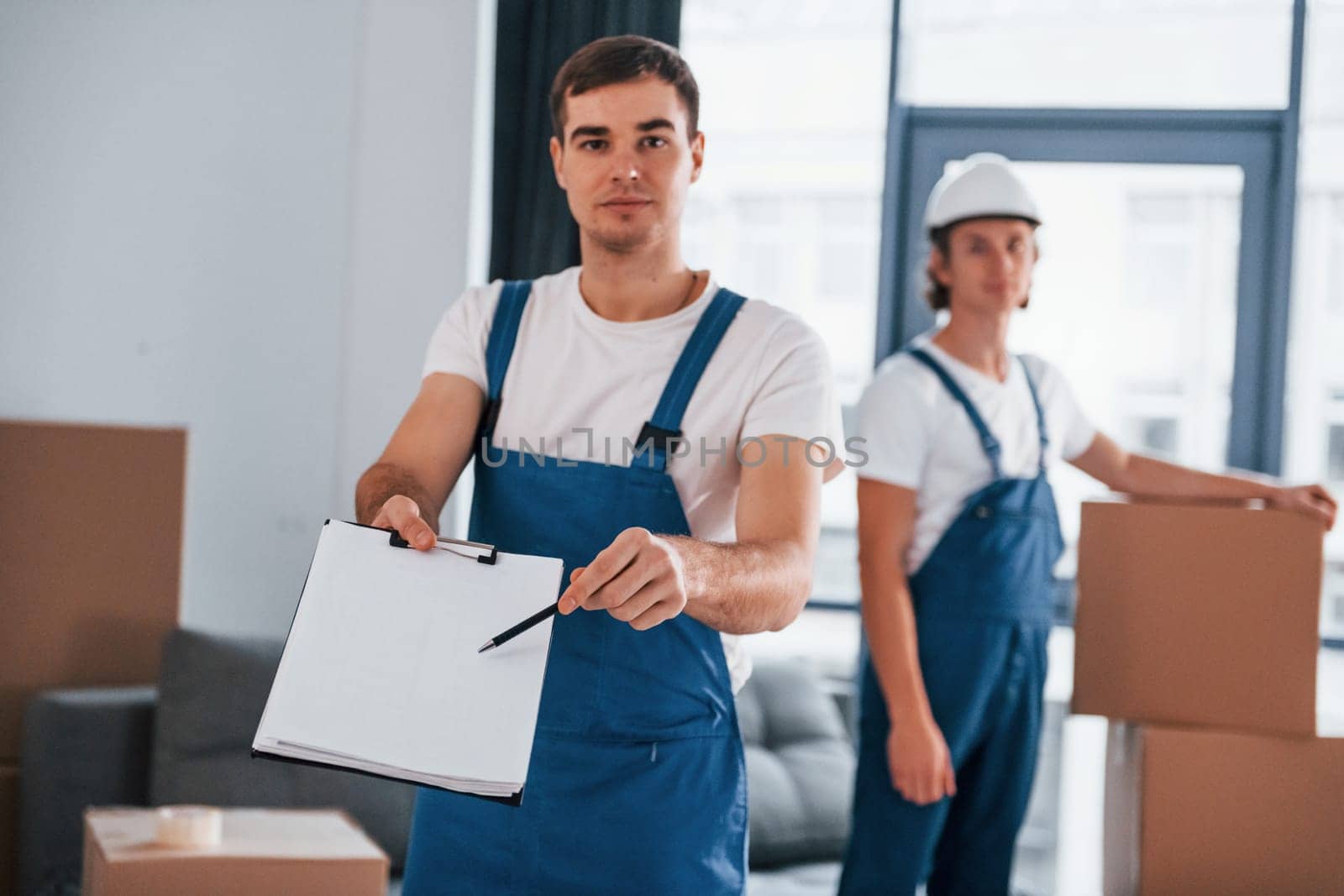 Document to sign. Two young movers in blue uniform working indoors in the room.