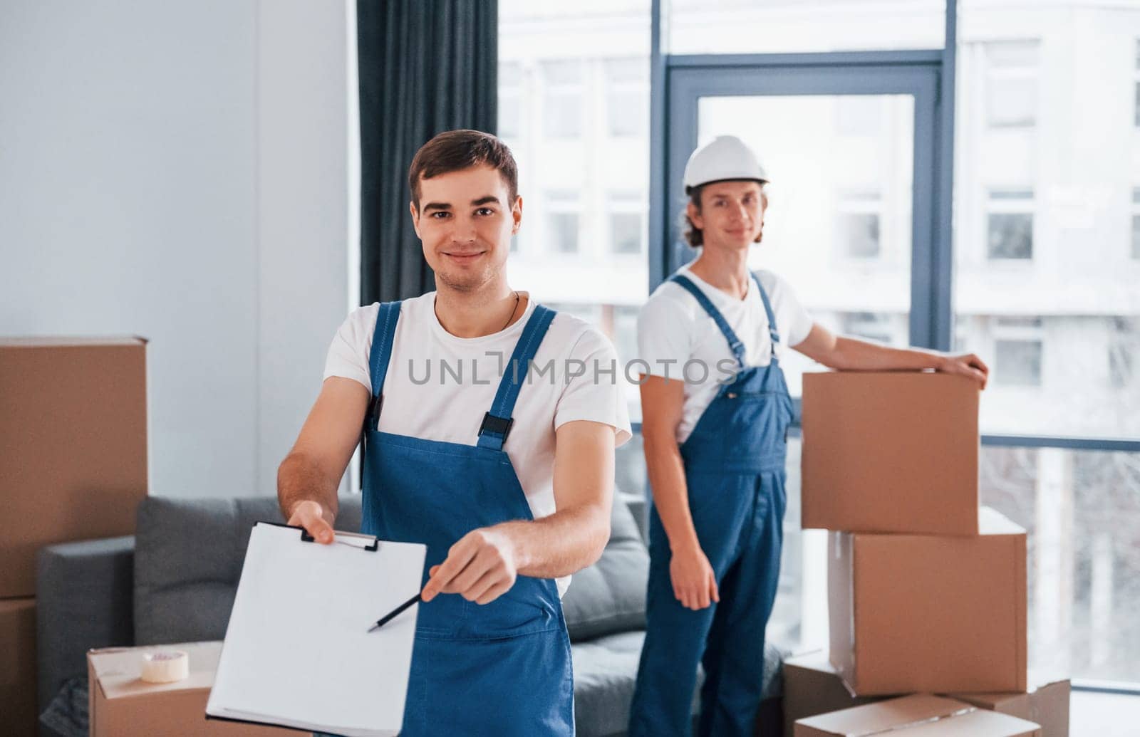 Document to sign. Two young movers in blue uniform working indoors in the room.