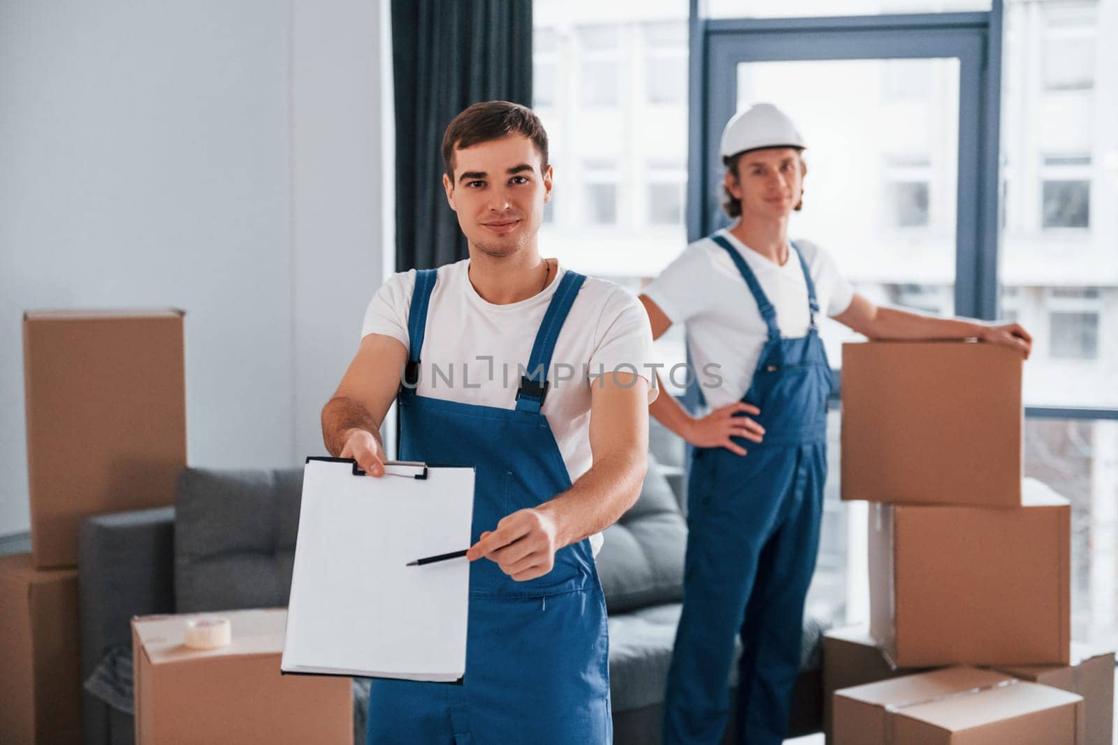 Document to sign. Two young movers in blue uniform working indoors in the room.
