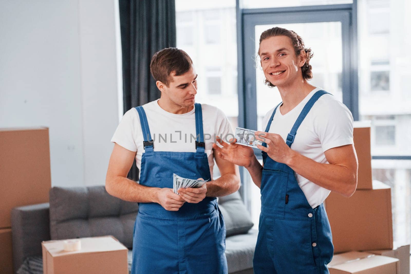 Holding money. Two young movers in blue uniform working indoors in the room.