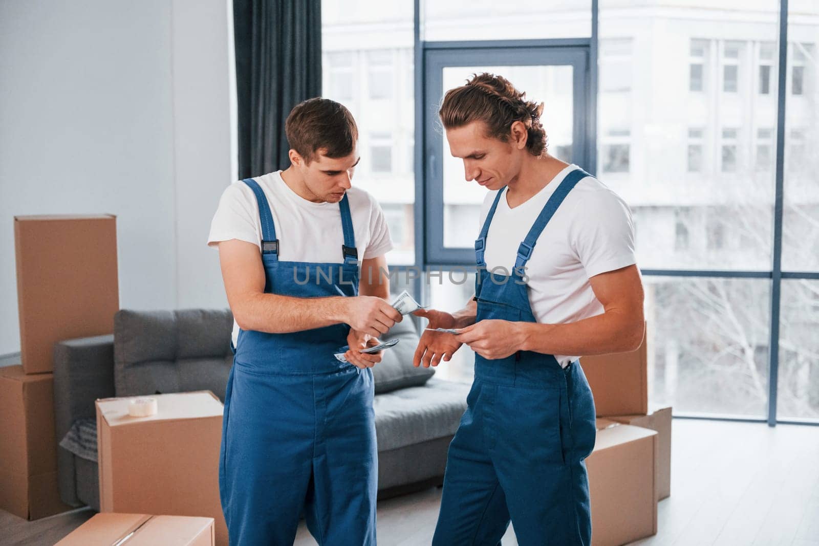 Holding money. Two young movers in blue uniform working indoors in the room.
