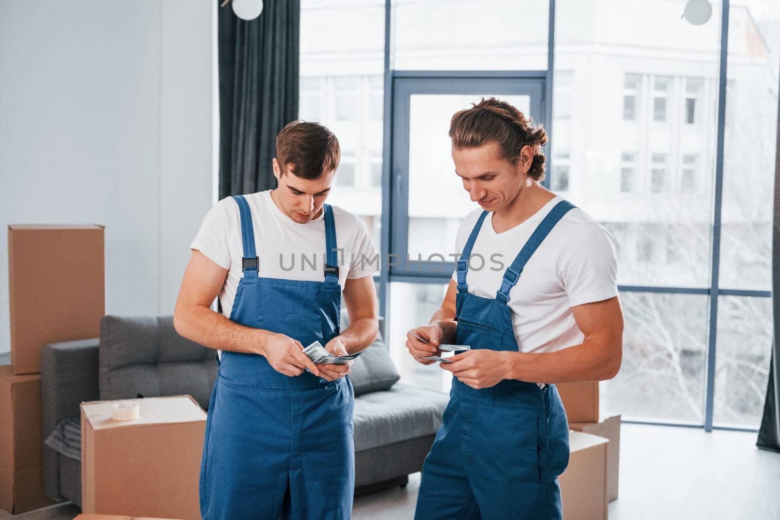 Holding money. Two young movers in blue uniform working indoors in the room.