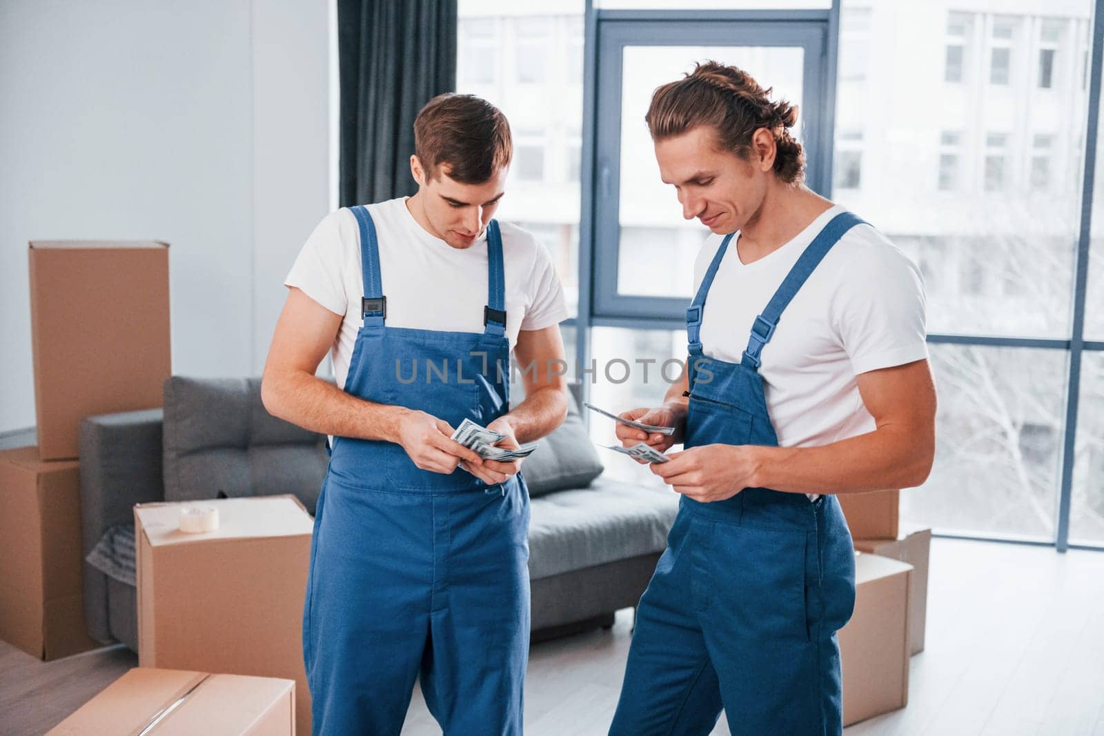 Holding money. Two young movers in blue uniform working indoors in the room.
