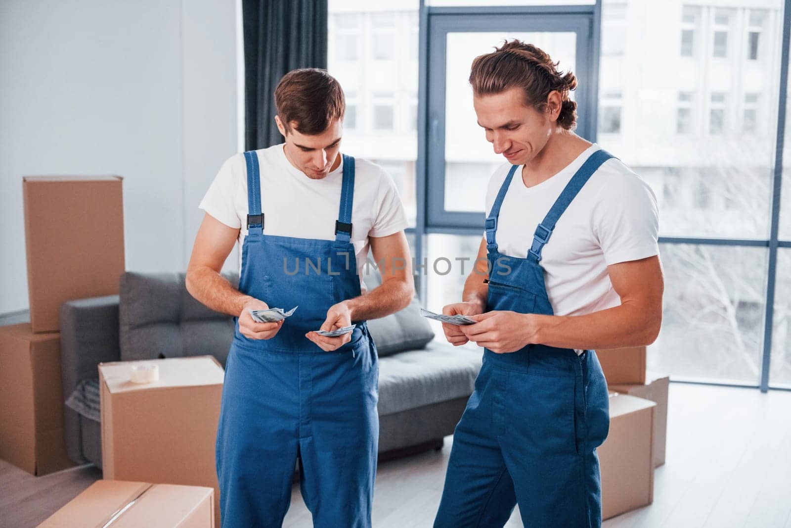 Holding money. Two young movers in blue uniform working indoors in the room.