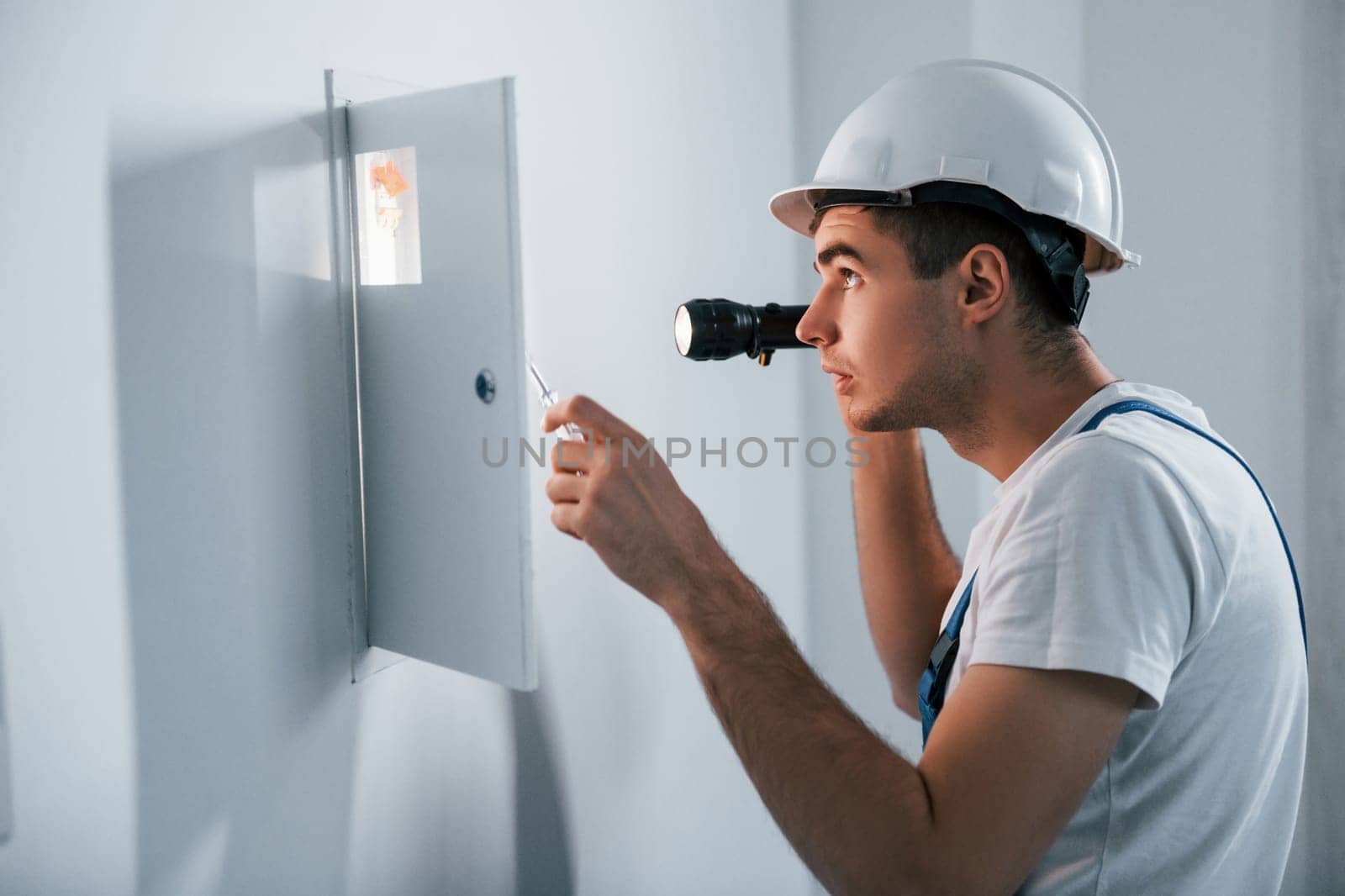Young male electrician in white hard hat works indoors in the room.