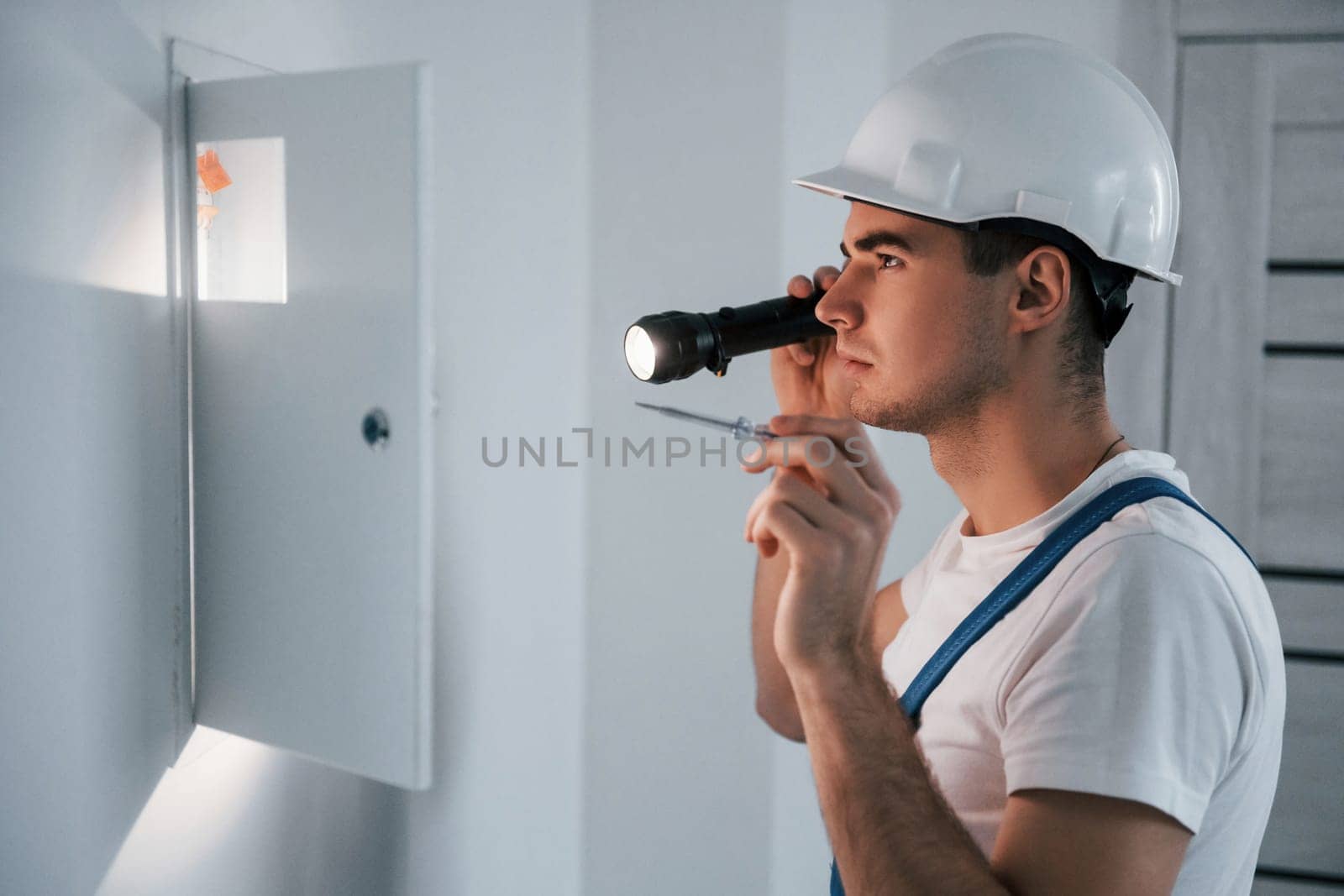Young male electrician in white hard hat works indoors in the room.