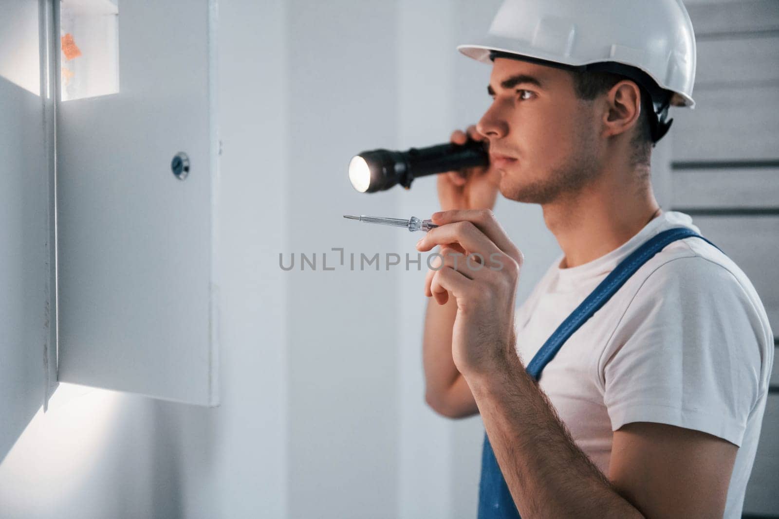 Young male electrician in white hard hat works indoors in the room.