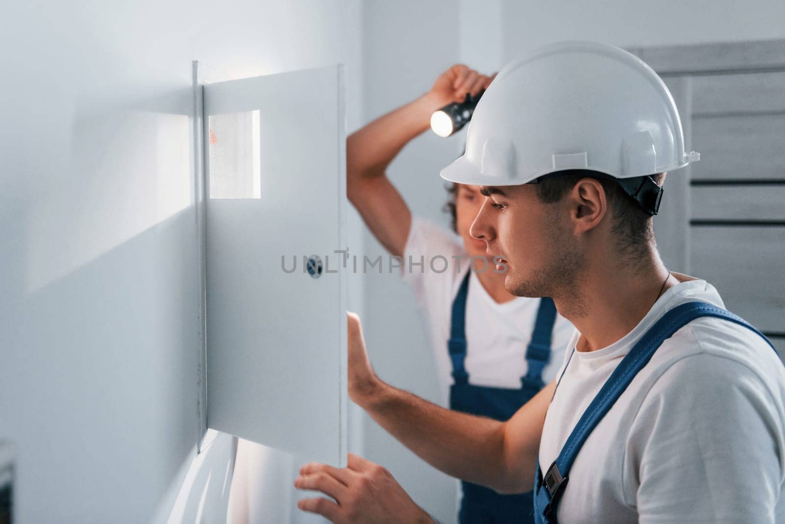 Two young male electricians works indoors together. Using flashlight.