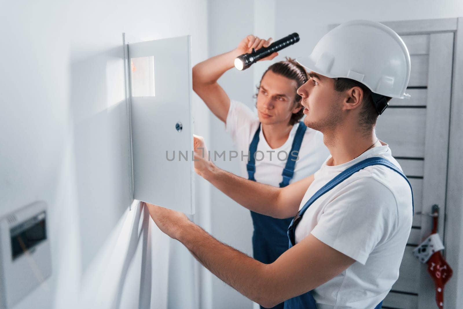Two young male electricians works indoors together. Using flashlight.