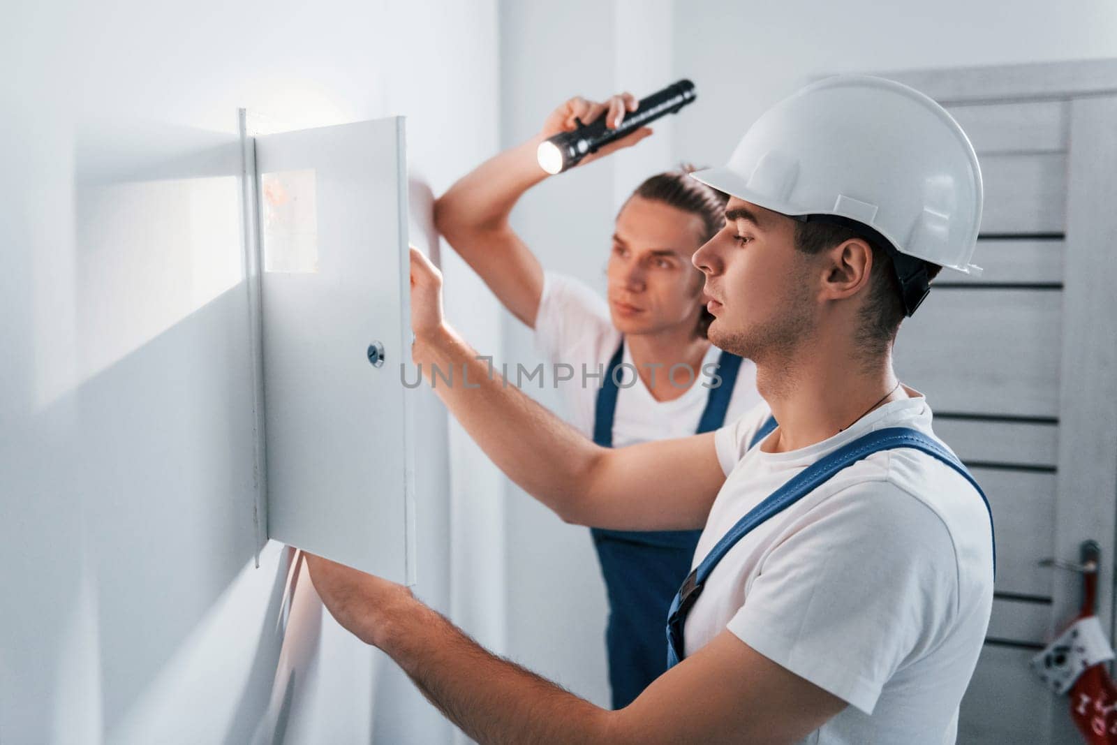 Two young male electricians works indoors together. Using flashlight.