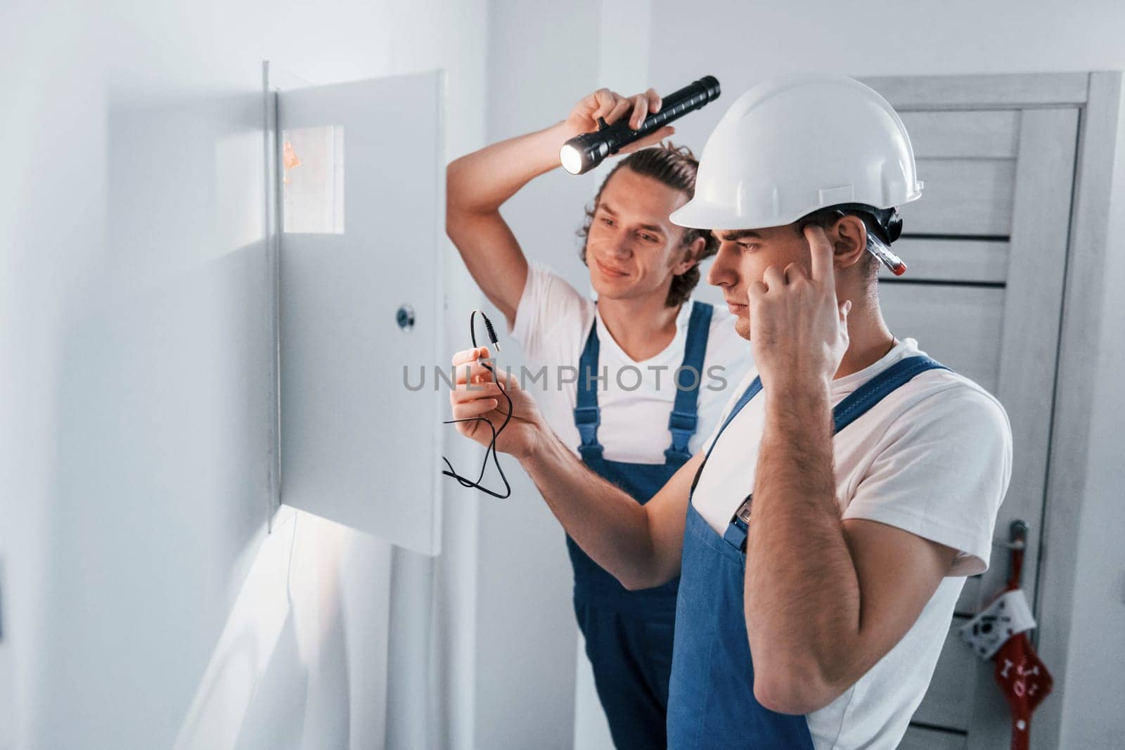 Two young male electricians works indoors together. Using flashlight.