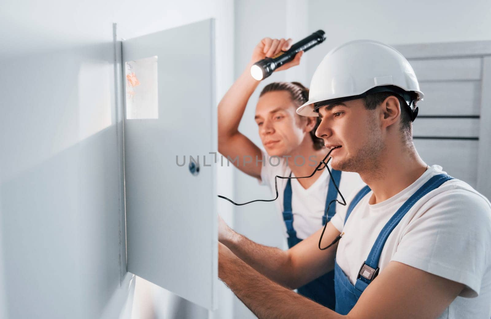 Two young male electricians works indoors together. Using flashlight.