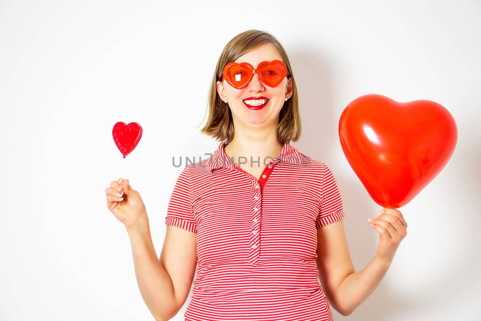 Portrait of the beautiful smiling woman in red sunglasses holding in her hands a red heart balloon and a red heart lollipop for Valentines Day. High quality photo