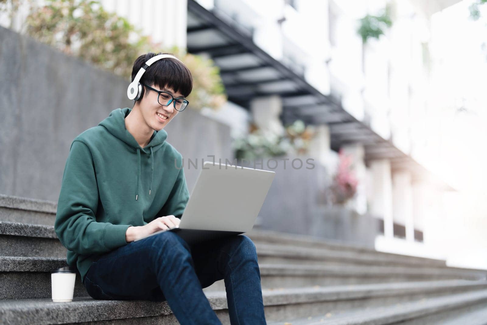 Smart Asian male college student wearing headphones, using laptop on campus outdoor stairs by wichayada