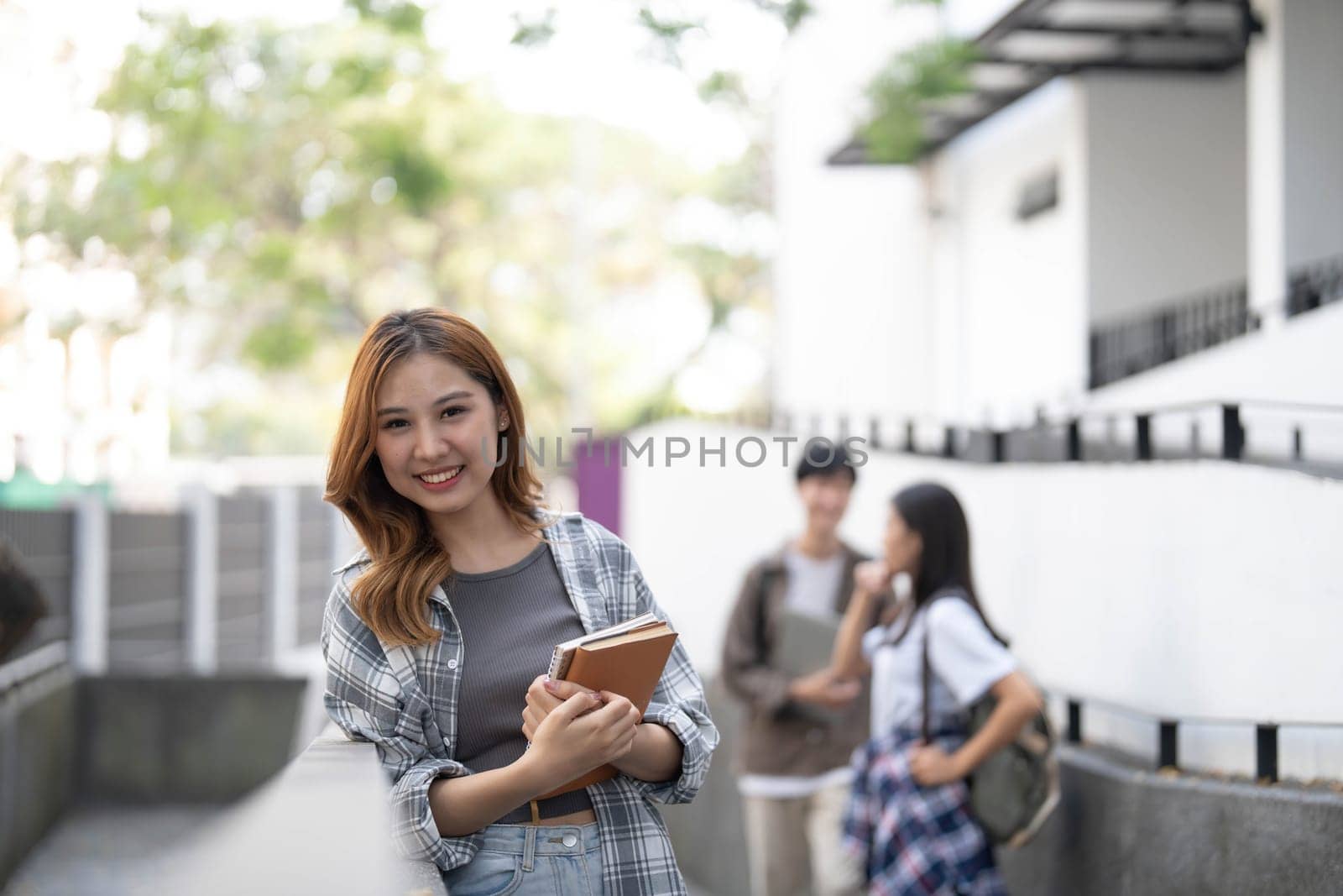 Cute asian girl student with hold a book and laptop near the campus against the background of a group of students. by wichayada