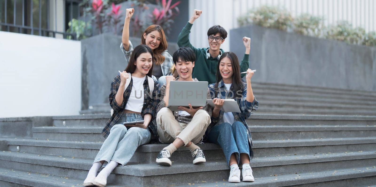 Group of cheerful Asian college students sitting on stairs, showing fists, celebrating triumph by wichayada