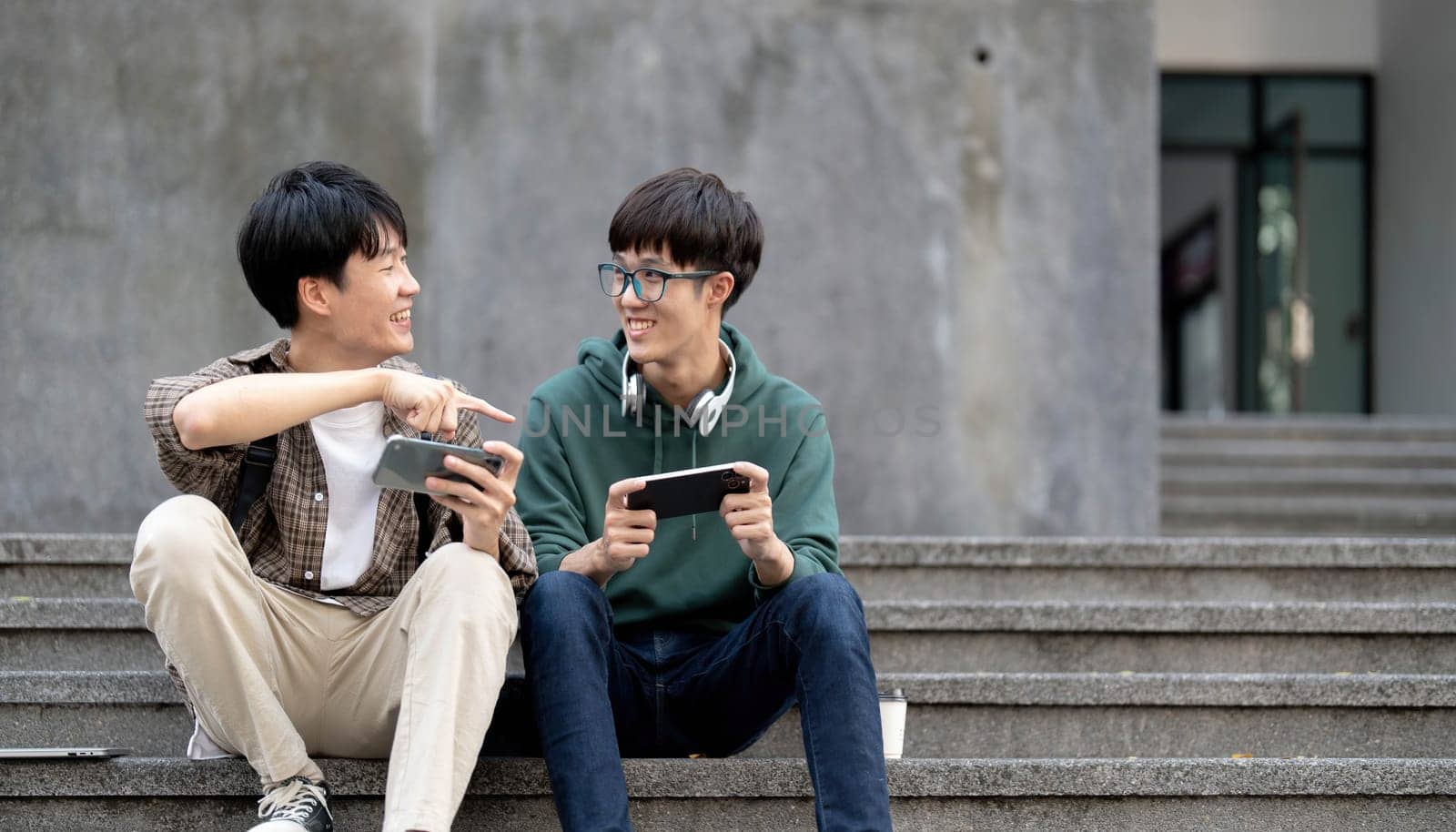 Happy Asian man college student playing mobile game on his smartphone with his friend by wichayada