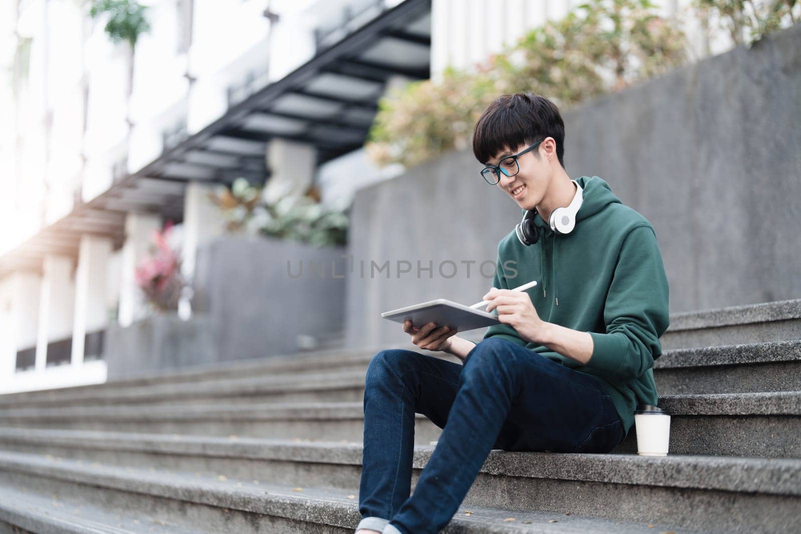 Smart Asian male college student wearing headphones, using laptop on campus outdoor stairs..