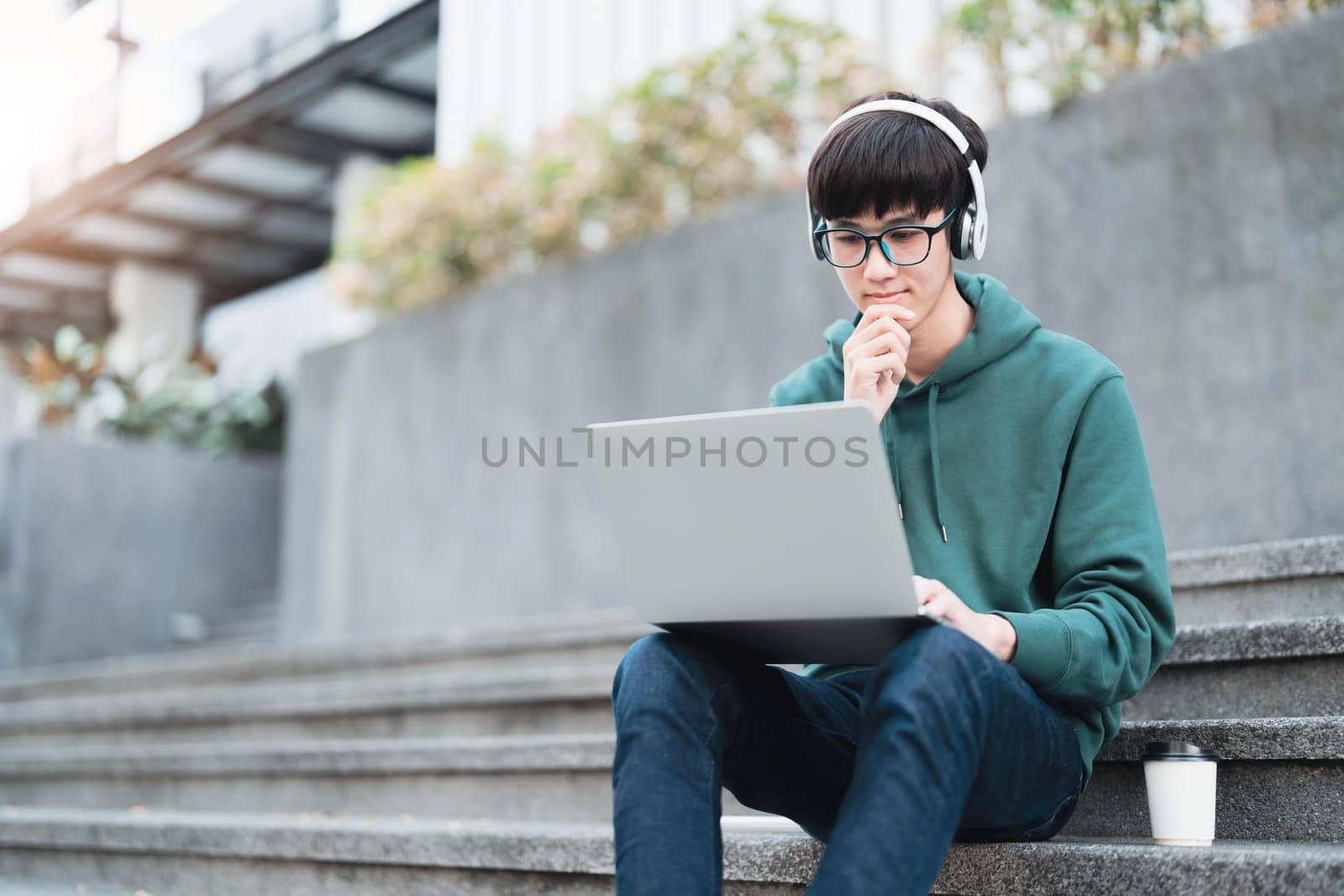 Smart Asian male college student wearing headphones, using laptop on campus outdoor stairs..