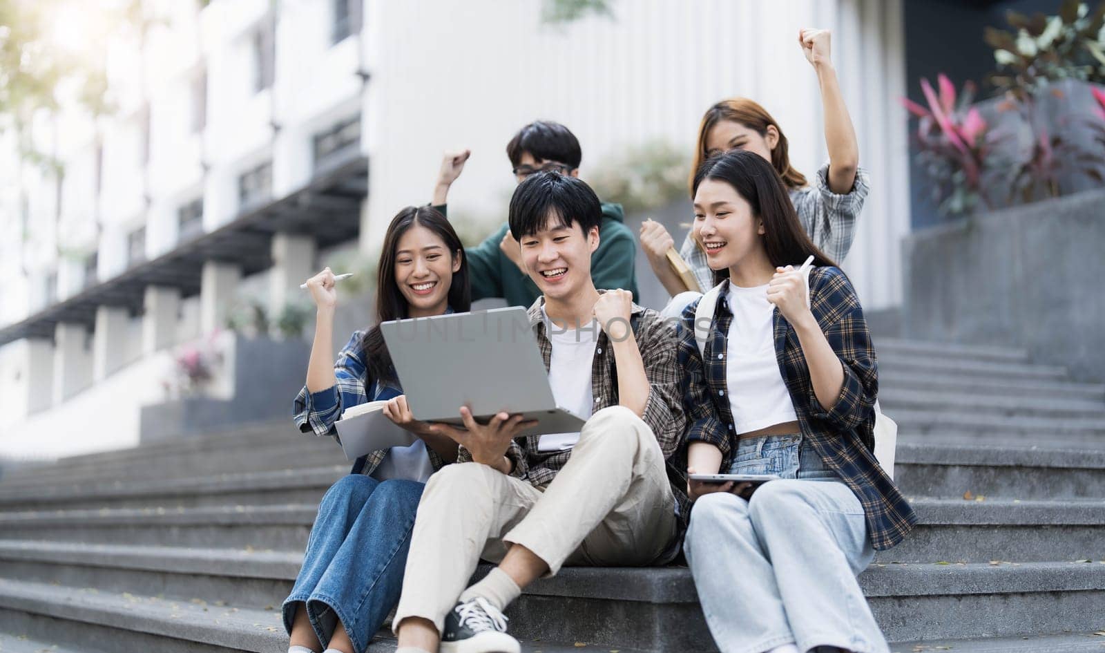 Group of cheerful Asian college students sitting on stairs, showing fists, celebrating triumph by wichayada