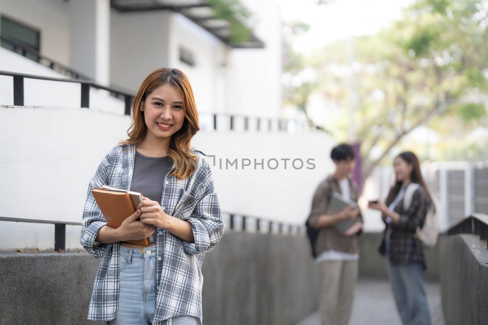 Cute asian girl student with hold a book and laptop near the campus against the background of a group of students. by wichayada