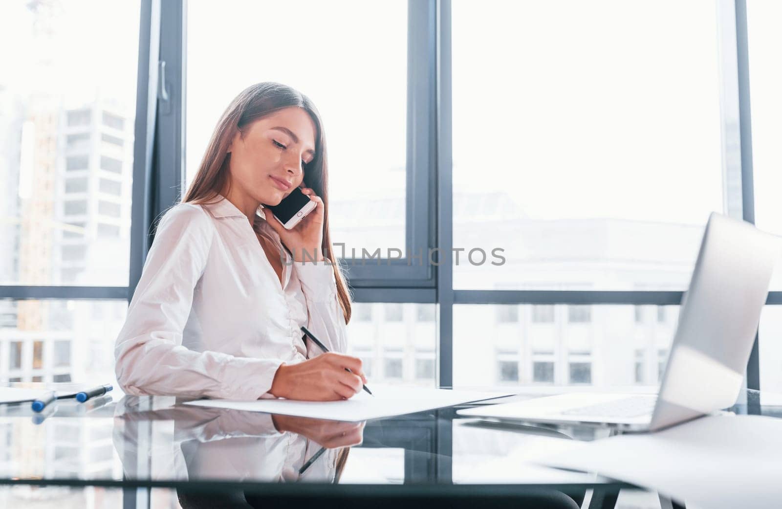Have conversation by the phone. Young woman in white formal clothes is indoors in the modern office.