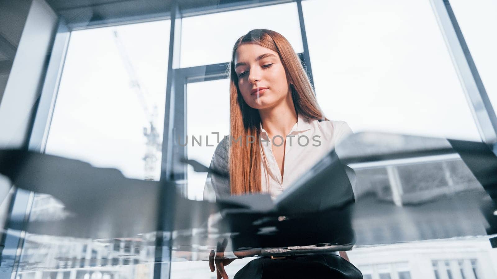Sits by the table. Young woman in white formal clothes is indoors in the modern office.