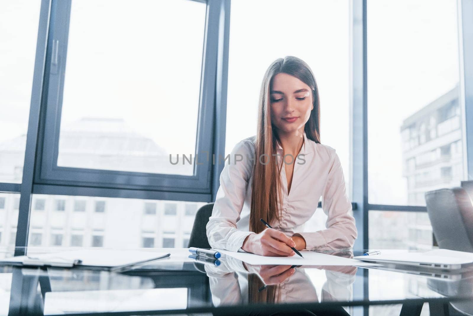 Sits by the table. Young woman in white formal clothes is indoors in the modern office.