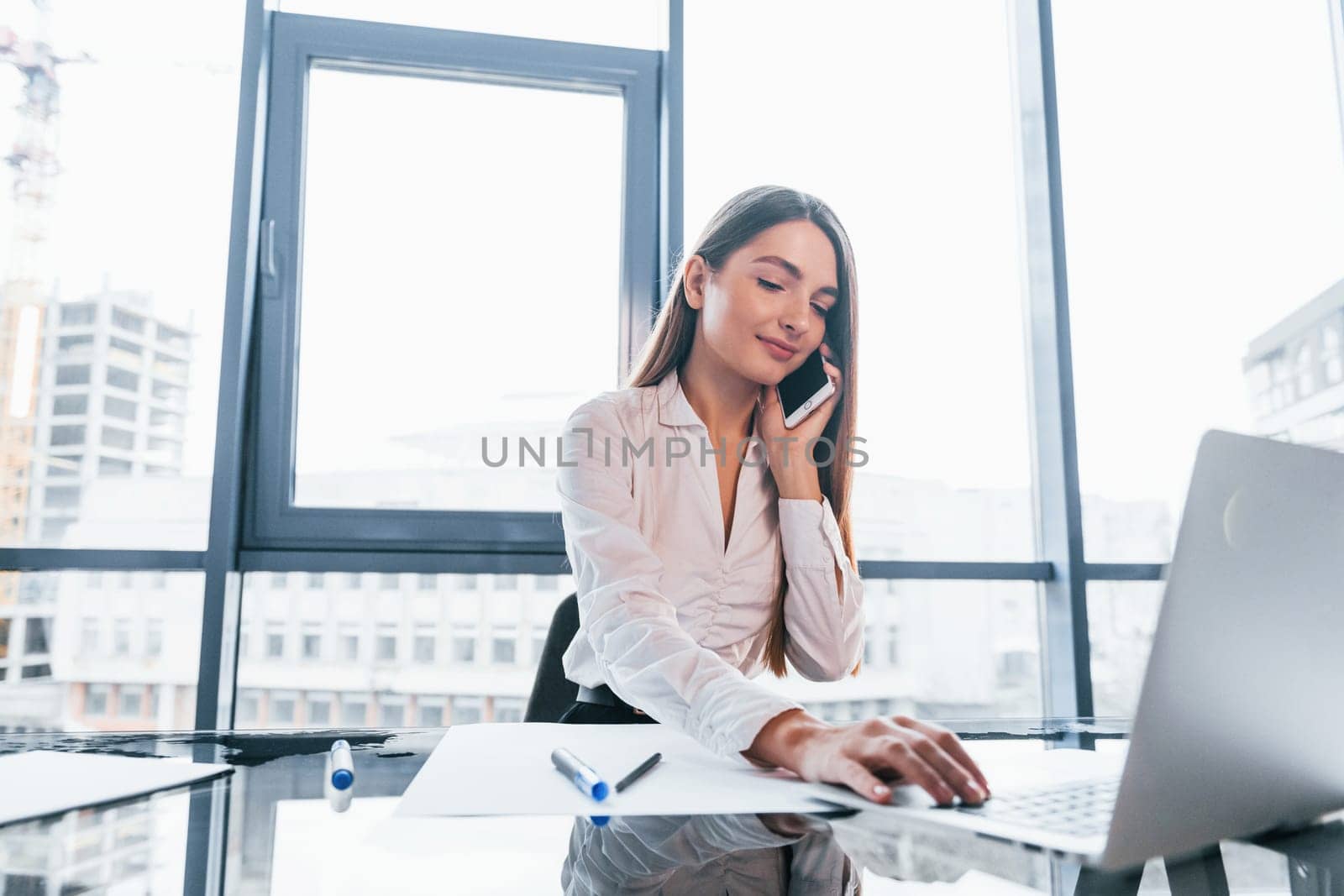 Sits by the table. Young woman in white formal clothes is indoors in the modern office.