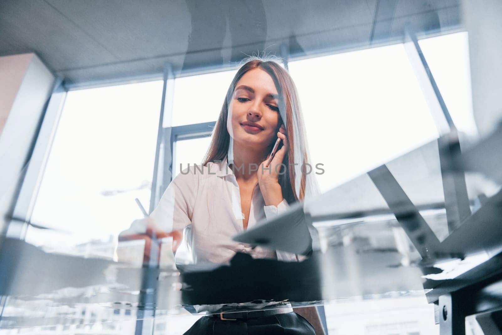 Have conversation by the phone. Young woman in white formal clothes is indoors in the modern office.