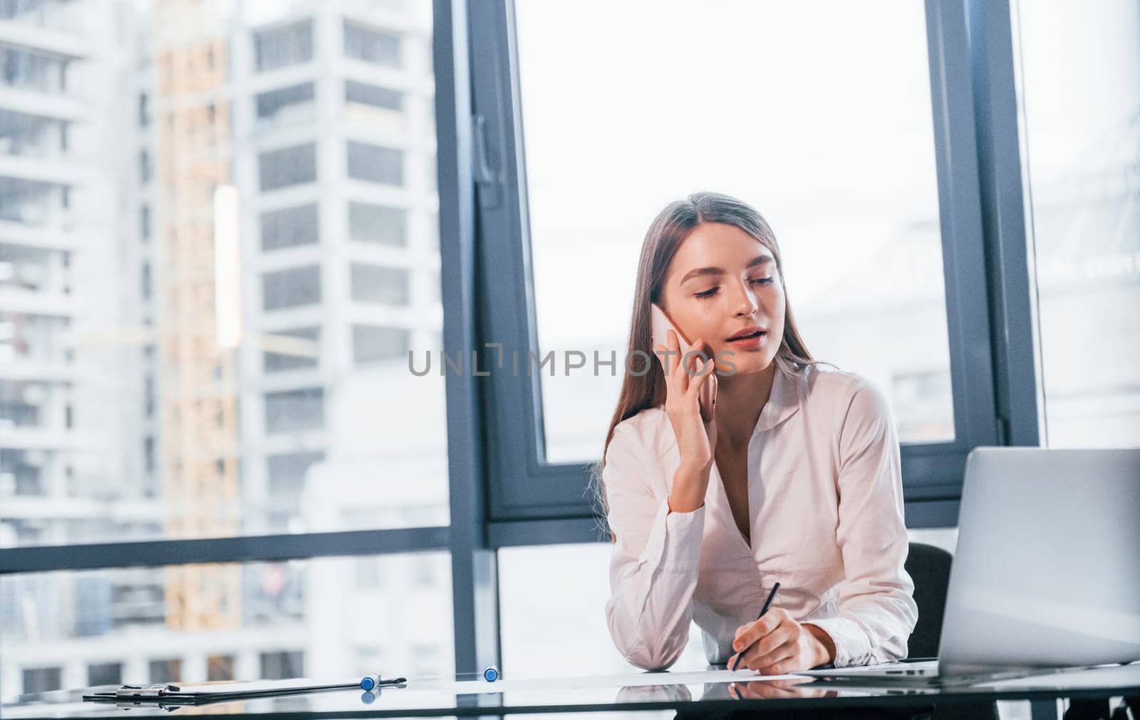 Have conversation by the phone. Young woman in white formal clothes is indoors in the modern office.