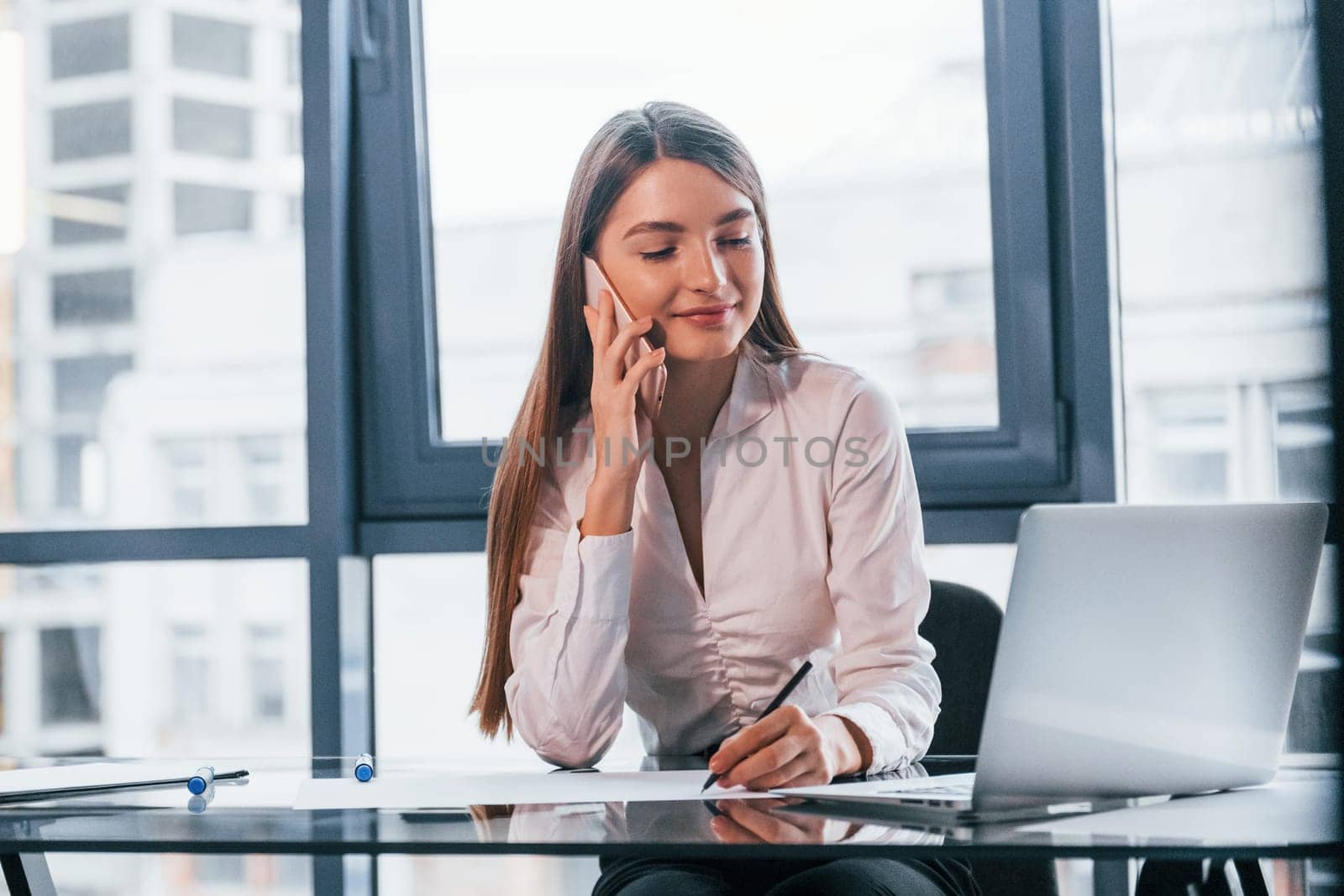 Have conversation by the phone. Young woman in white formal clothes is indoors in the modern office.
