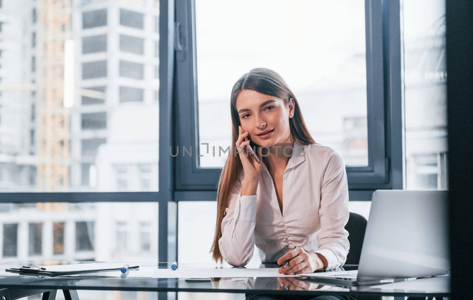 Have conversation by the phone. Young woman in white formal clothes is indoors in the modern office.