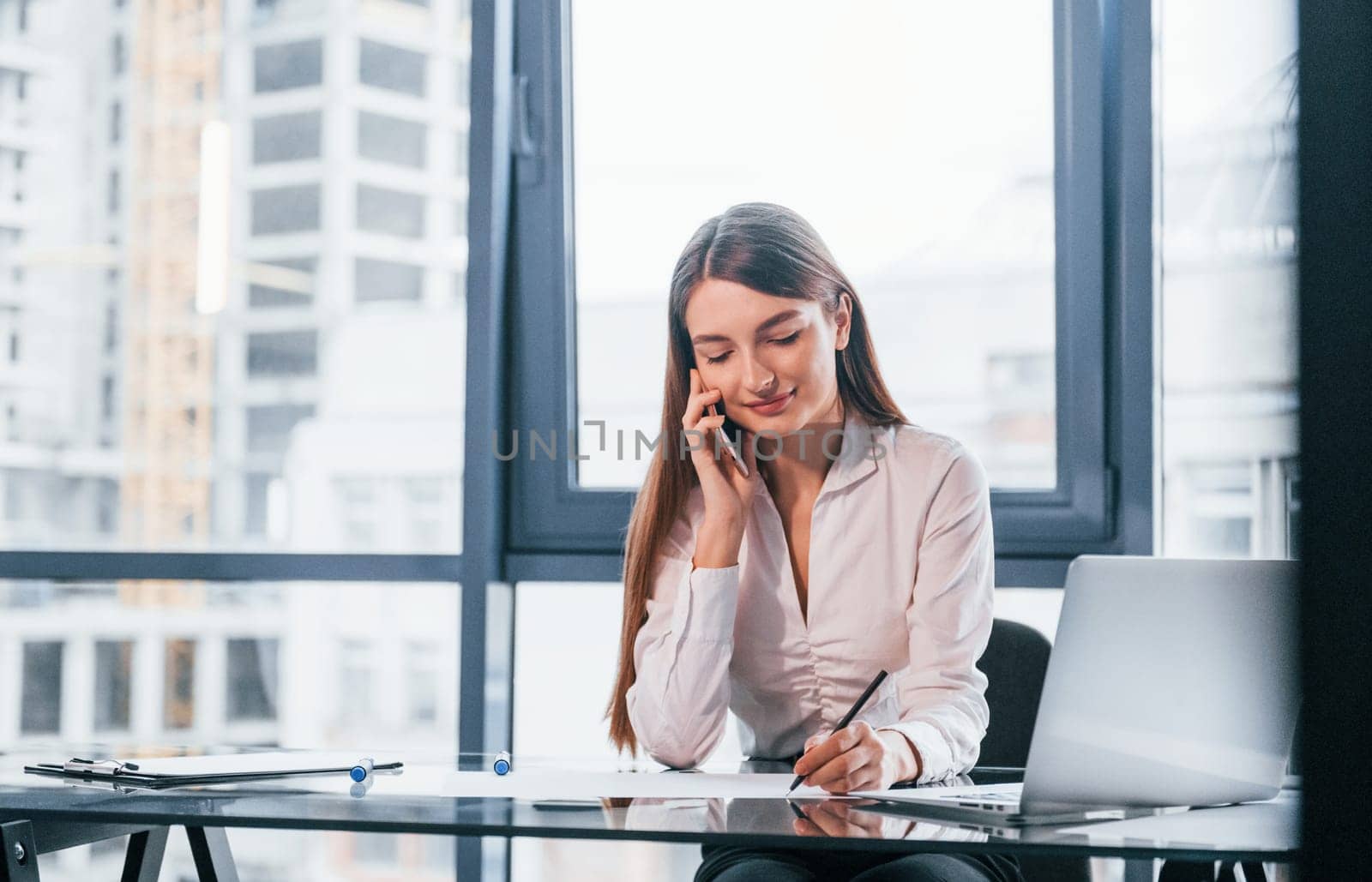 Have conversation by the phone. Young woman in white formal clothes is indoors in the modern office.