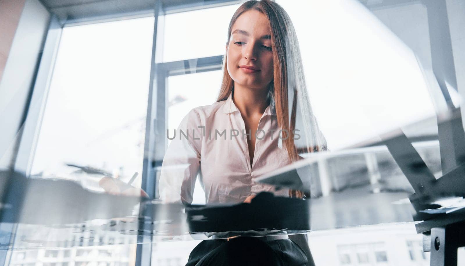 Sits by the table. Young woman in white formal clothes is indoors in the modern office.