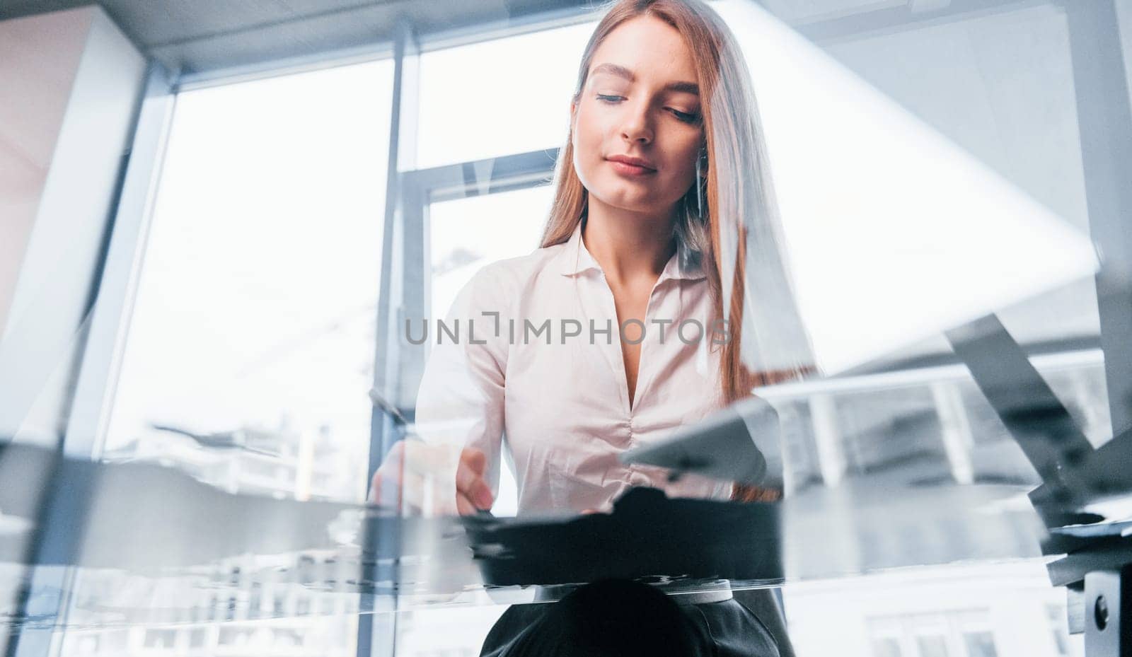 Sits by the table. Young woman in white formal clothes is indoors in the modern office.