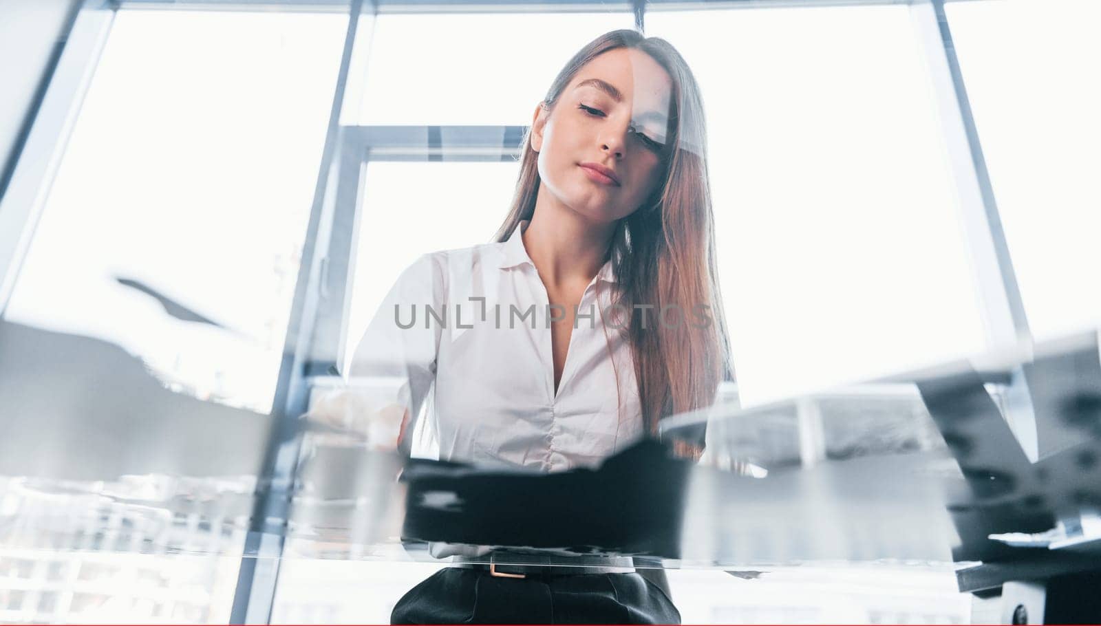 Sits by the table. Young woman in white formal clothes is indoors in the modern office.