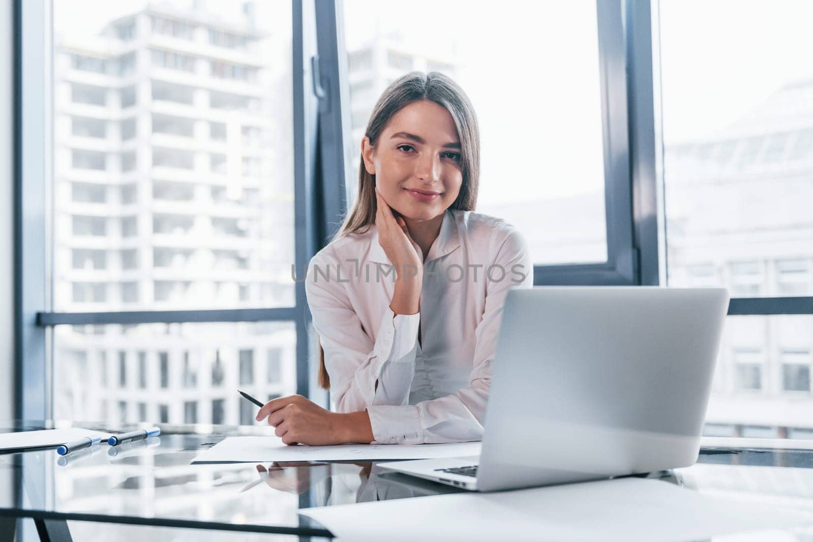 Young woman in white formal clothes is indoors in the modern office.