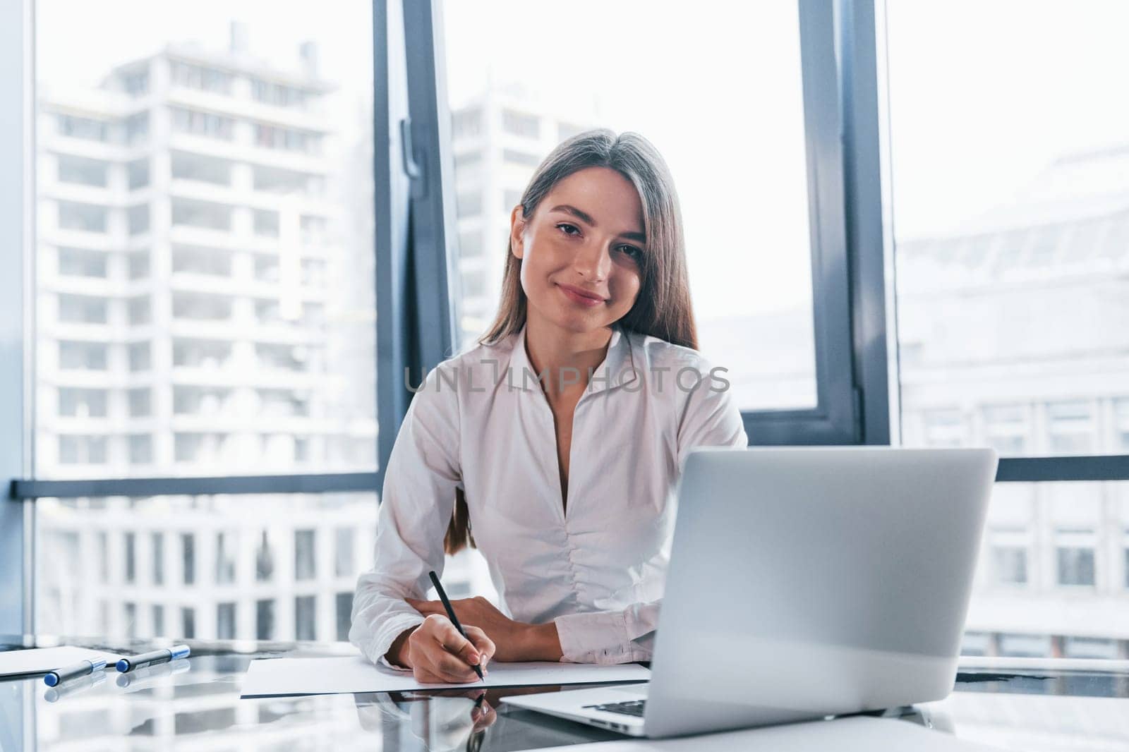 Young woman in white formal clothes is indoors in the modern office.