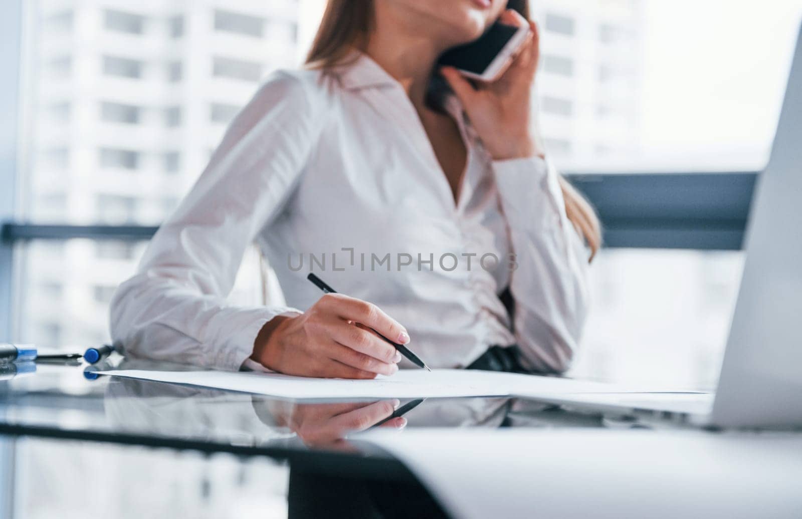 Have conversation by the phone. Young woman in white formal clothes is indoors in the modern office.
