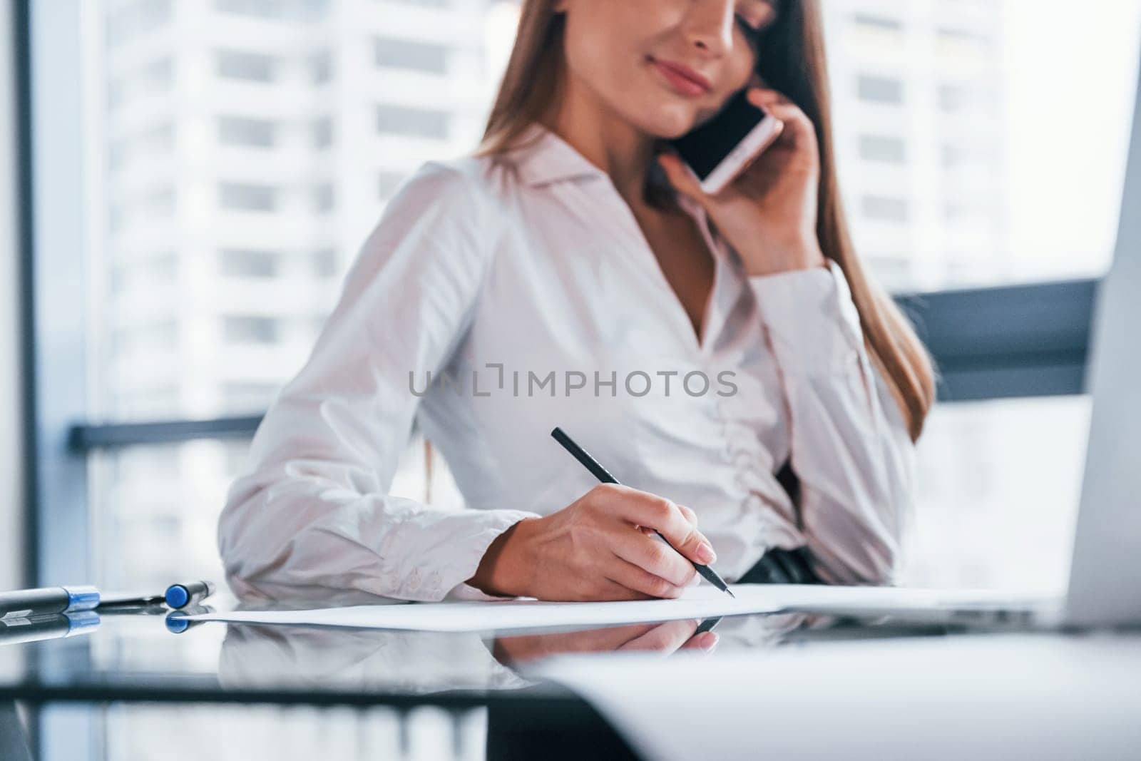 Have conversation by the phone. Young woman in white formal clothes is indoors in the modern office.
