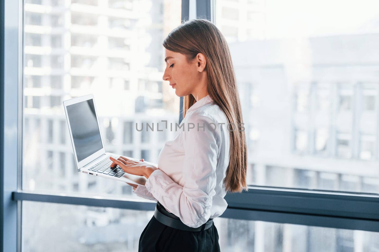 Standing with laptop. Young woman in white formal clothes is indoors in the modern office.